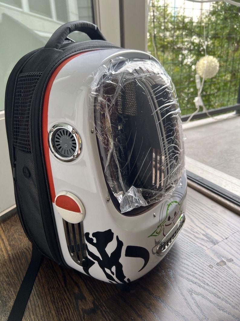 Puede incluir: Una mochila porta mascotas blanca y negra con una ventana de pl&aacute;stico transparente. La mochila tiene una franja roja y un ventilador de ventilaci&oacute;n plateado. La mochila tiene un gr&aacute;fico en blanco y negro de un personaje de Star Wars en el lateral.