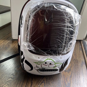 Puede incluir: Una mochila blanca y negra con una burbuja de pl&aacute;stico transparente en la parte delantera. La burbuja tiene un peque&ntilde;o personaje verde en su interior. La mochila tiene una correa negra y un asa negra.