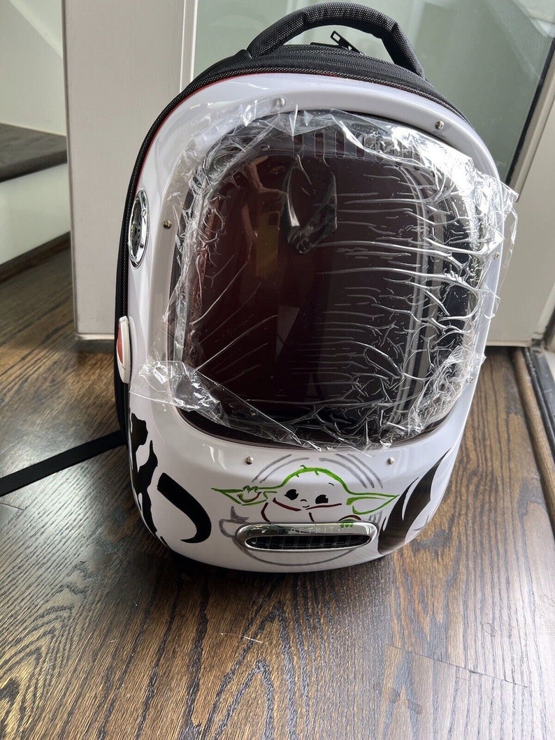 Puede incluir: Una mochila blanca y negra con una burbuja de pl&aacute;stico transparente en la parte delantera. La burbuja tiene un peque&ntilde;o personaje verde en su interior. La mochila tiene una correa negra y un asa negra.