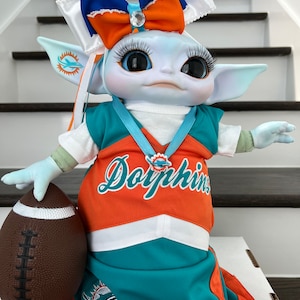 May include: A blue and orange plush toy dressed as a Miami Dolphins cheerleader, holding a brown football. The toy has large eyes, long eyelashes, and a white bow with a rhinestone in the centre.