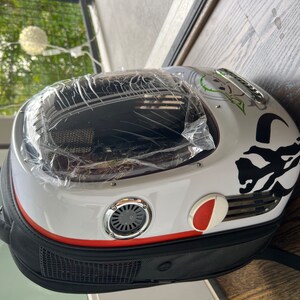 Puede incluir: Una mochila porta mascotas blanca y negra con una ventana de pl&aacute;stico transparente. La mochila tiene una franja roja y un dise&ntilde;o gr&aacute;fico en blanco y negro. La mochila tiene un asa negra y una correa negra.