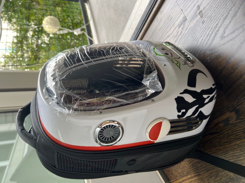 Puede incluir: Una mochila porta mascotas blanca y negra con una ventana de pl&aacute;stico transparente. La mochila tiene una franja roja y un dise&ntilde;o gr&aacute;fico en blanco y negro. La mochila tiene un asa negra y una correa negra.