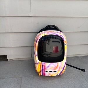 May include: A colorful, abstract patterned pet carrier backpack with a clear plastic window. The backpack has a black handle and a black strap.