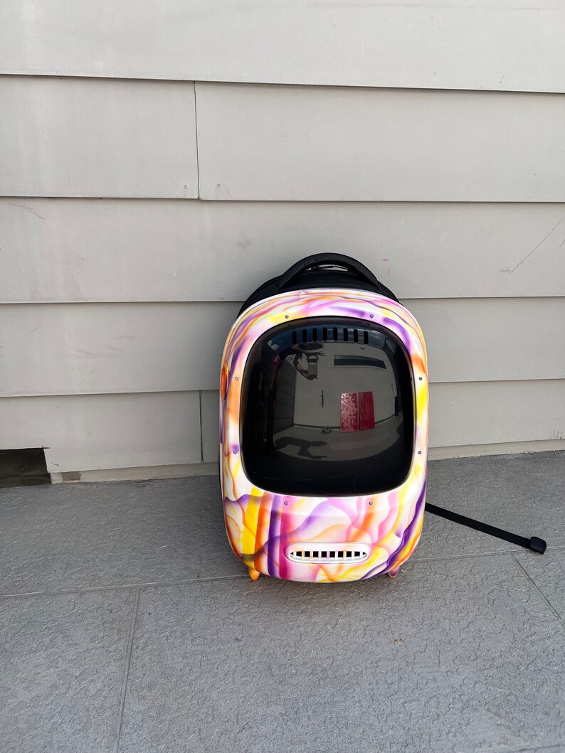 May include: A colorful, abstract patterned pet carrier backpack with a clear plastic window. The backpack has a black handle and a black strap.