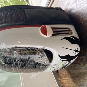 Puede incluir: Mochila portamascotas blanca y negra con un bot&oacute;n rojo y un s&iacute;mbolo de la Alianza Rebelde de Star Wars en blanco y negro. La mochila tiene una parte delantera de pl&aacute;stico transparente.