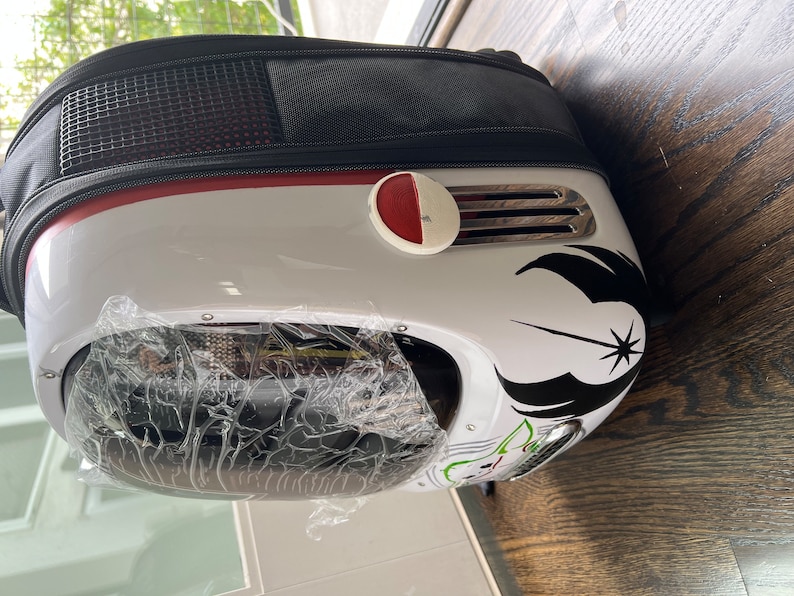 Puede incluir: Mochila portamascotas blanca y negra con un bot&oacute;n rojo y un s&iacute;mbolo de la Alianza Rebelde de Star Wars en blanco y negro. La mochila tiene una parte delantera de pl&aacute;stico transparente.