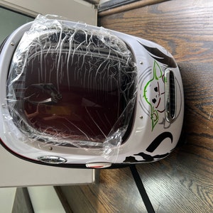 Puede incluir: Mochila blanca con una ventana de pl&aacute;stico transparente en la parte delantera. La mochila tiene un gr&aacute;fico verde y negro de un personaje de la franquicia de Star Wars, "The Mandalorian".