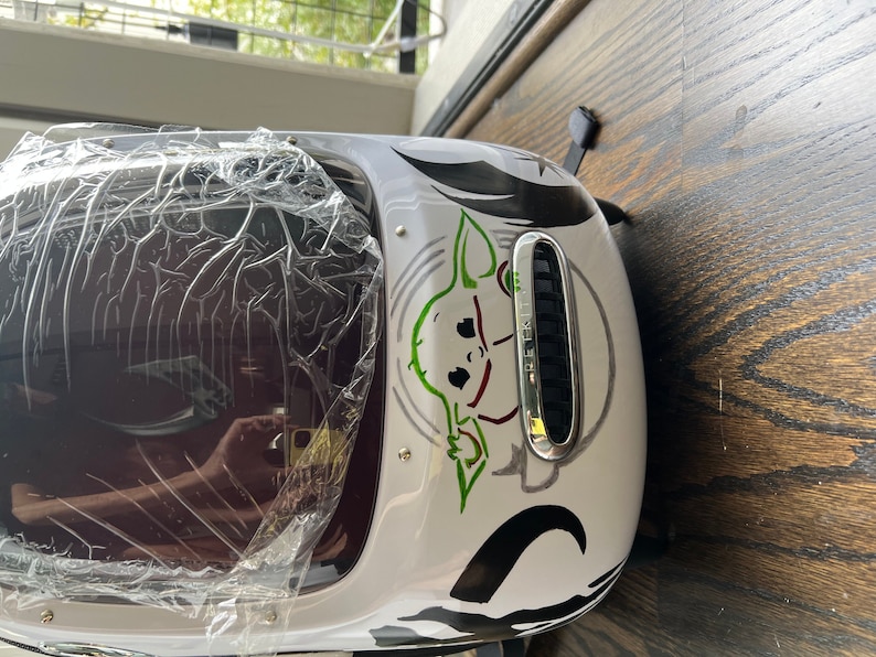 Puede incluir: Mochila blanca con un gr&aacute;fico negro y verde de un personaje de la franquicia Star Wars. El personaje es un extraterrestre verde con orejas grandes y una expresi&oacute;n traviesa. La mochila tiene una ventana de pl&aacute;stico transparente en la parte delantera.