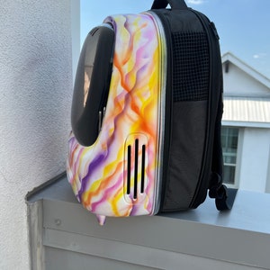 May include: A white and purple pet carrier backpack with a black mesh pocket on the side. The carrier has a clear plastic bubble on the front and a ventilation system on the side.