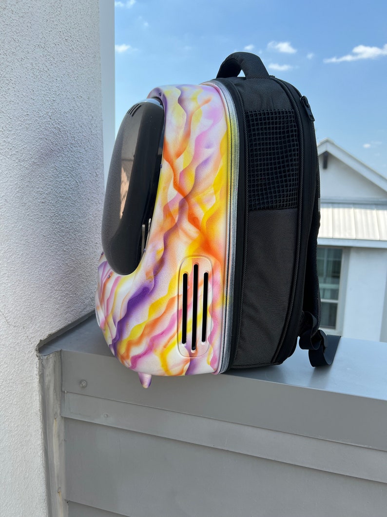 May include: A white and purple pet carrier backpack with a black mesh pocket on the side. The carrier has a clear plastic bubble on the front and a ventilation system on the side.