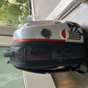 Puede incluir: Una mochila porta mascotas blanca y negra con una franja roja y una ventana de malla. La mochila tiene un asa negra y una correa negra.