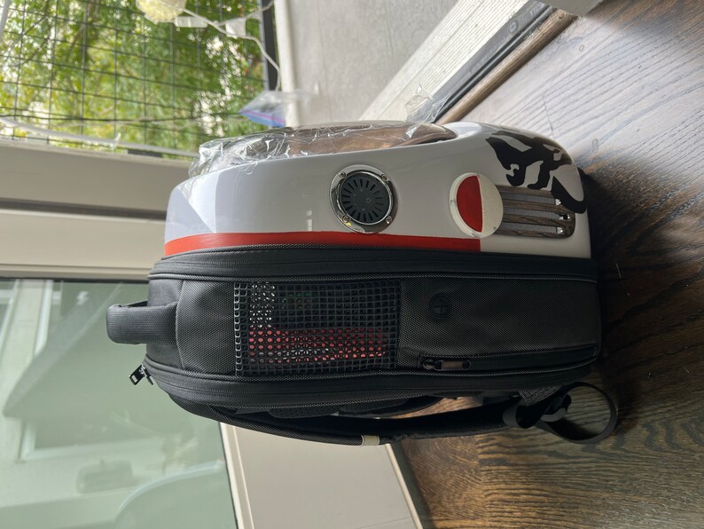 Puede incluir: Una mochila porta mascotas blanca y negra con una franja roja y una ventana de malla. La mochila tiene un asa negra y una correa negra.