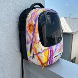 May include: A purple, yellow, and orange patterned pet carrier backpack with a clear bubble for the pet to see out of. The backpack has a black strap and a black mesh panel on the side.
