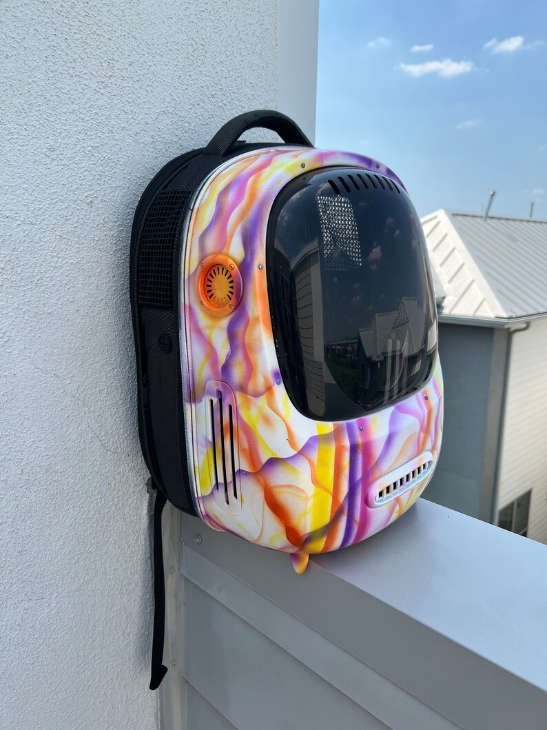 May include: A purple, yellow, and orange patterned pet carrier backpack with a clear bubble for the pet to see out of. The backpack has a black strap and a black mesh panel on the side.