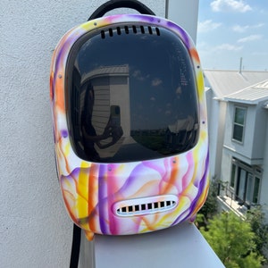 May include: A colorful, tie-dye patterned pet carrier backpack with a clear bubble for visibility. The backpack has a black strap and a black handle.