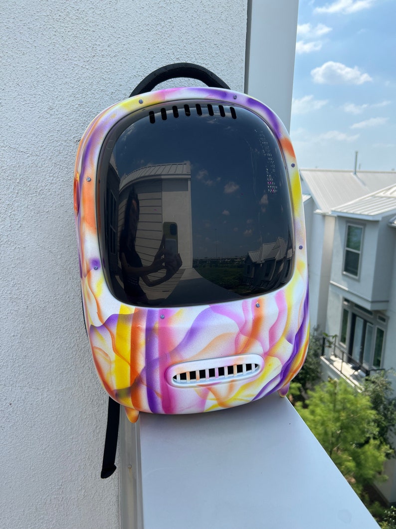 May include: A colorful, tie-dye patterned pet carrier backpack with a clear bubble for visibility. The backpack has a black strap and a black handle.