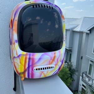 May include: A white, purple, and yellow patterned backpack with a clear plastic bubble on the front. The backpack has a black strap and a small vent on the front.