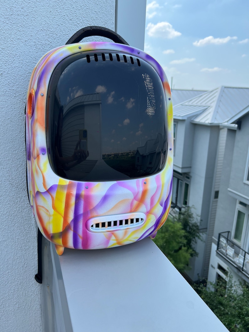 May include: A white, purple, and yellow patterned backpack with a clear plastic bubble on the front. The backpack has a black strap and a small vent on the front.