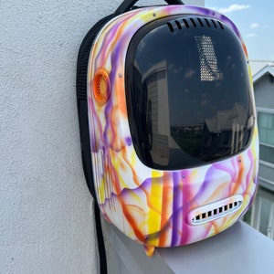 May include: A white backpack with a colorful abstract design in shades of purple, orange, and yellow. The backpack has a large clear bubble-like window on the front and a black strap.