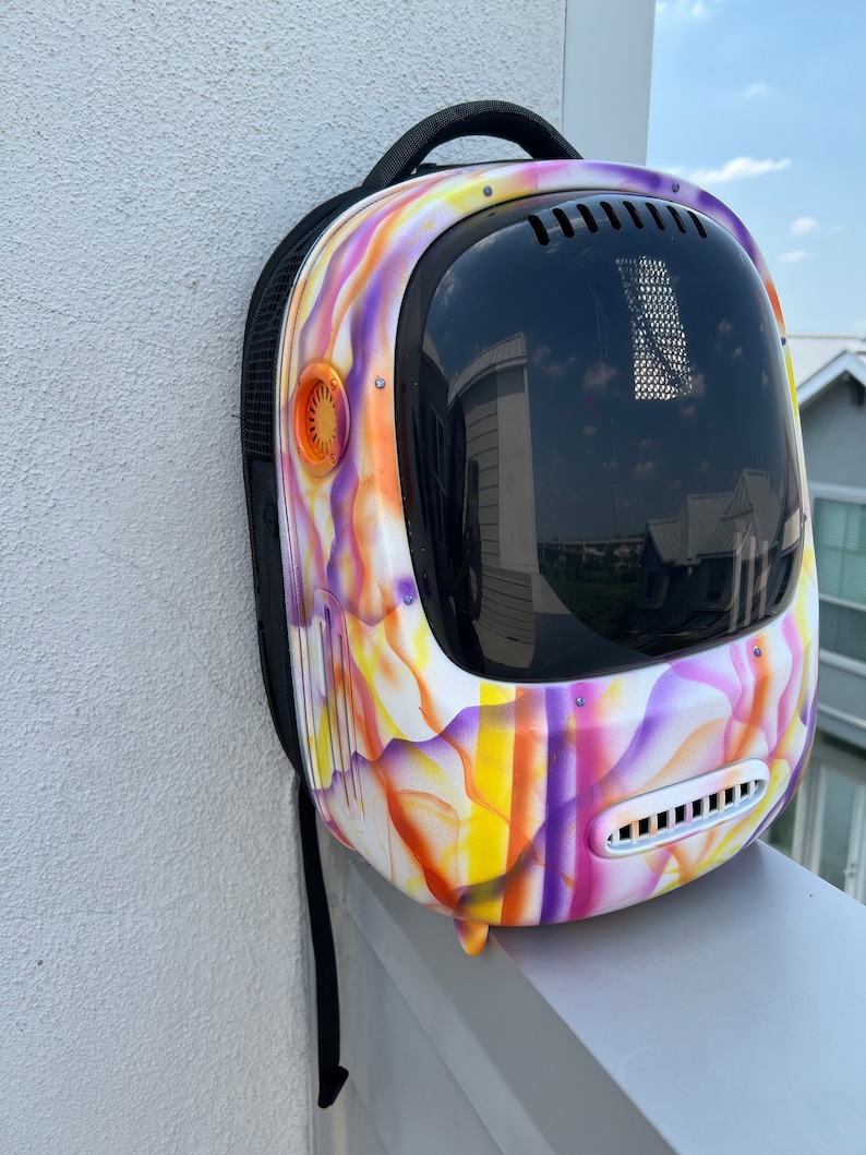 May include: A white backpack with a colorful abstract design in shades of purple, orange, and yellow. The backpack has a large clear bubble-like window on the front and a black strap.