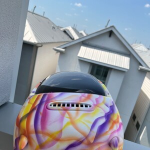 May include: A white helmet with a colorful abstract design featuring purple, yellow, and pink swirls. The helmet has a black visor and a white vent.