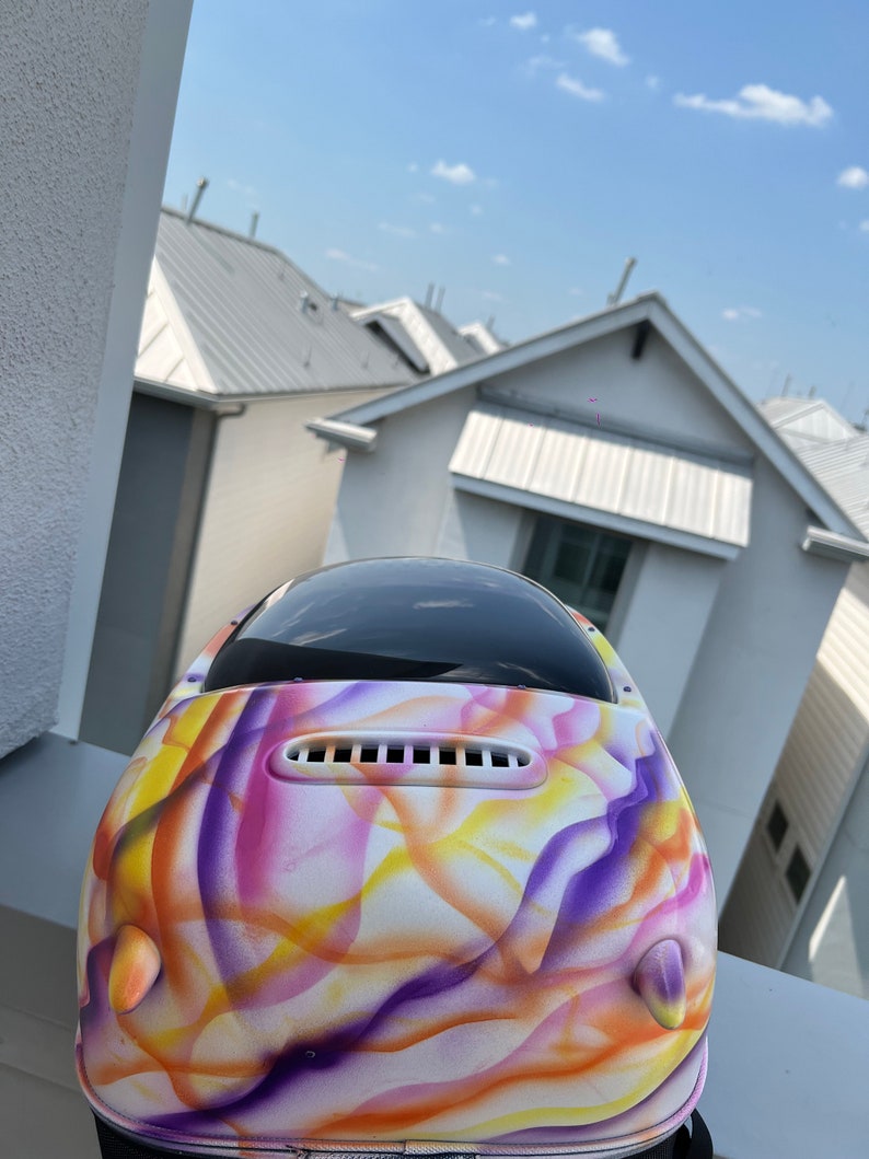May include: A white helmet with a colorful abstract design featuring purple, yellow, and pink swirls. The helmet has a black visor and a white vent.