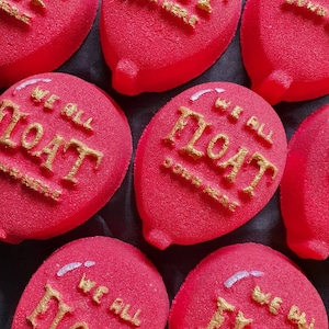 May include: Close-up of several bright red, balloon-shaped objects with gold lettering that reads "WE ALL FLOAT DOWN HERE". The objects have a textured surface and a small tail. The background is black.