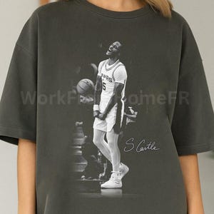 May include: Dark gray oversized t-shirt featuring a black and white image of a basketball player in action. The player is wearing a white uniform with the number 5. The name "S Castle" is written in cursive. The text "WorkFromHomeFR" is also visible.