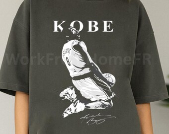 May include: Dark gray t-shirt featuring a black and white image of a basketball player in action, with the name "KOBE" above the image. The player's jersey number is 8, and the shirt includes a signature.