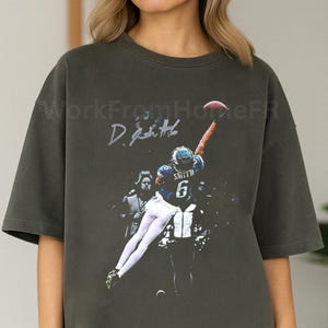 May include: Dark grey t-shirt featuring a graphic of a football player in action, reaching for a football. The player is wearing a blue and white uniform with the number 6. The shirt also has the text "WorkFromHomeFR" and a signature.