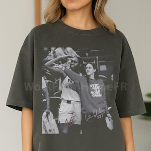 May include: Dark grey t-shirt featuring a black and white image of two women. One is raising her arm, and the other is holding a paper. The text "Every Home Game" is visible on the shirt. The shirt has a relaxed fit.