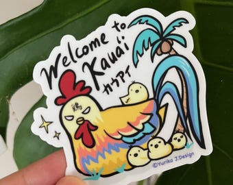 以下が含まれることがあります： 赤い鶏冠と黄色いひよこを持つ漫画の雄鶏が描かれたカラフルなステッカー。雄鶏はヤシの木と波に囲まれています。テキスト「Welcome to Kauai! Aloha!」は黒い筆記体で書かれています。