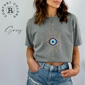 May include: A grey t-shirt with a gold evil eye design. The eye is blue with a white pupil and a black outline. The design is surrounded by gold rays.