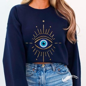 May include: A navy blue cropped sweatshirt with a gold evil eye design. The eye is blue with a white pupil and is surrounded by gold rays.