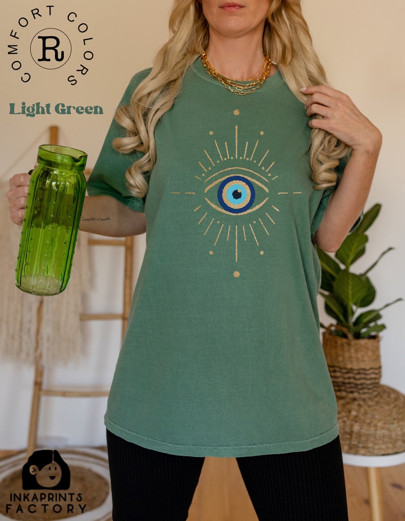 May include: A light green t-shirt with a gold glitter evil eye design. The shirt is a relaxed fit and has a crew neck.