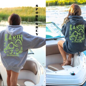 May include: Two women wearing hooded sweatshirts with the text "Earth is Home to Everyone" and a graphic of a smiling flower and rainbow. The sweatshirt on the left is sport grey and the sweatshirt on the right is dark heather grey.