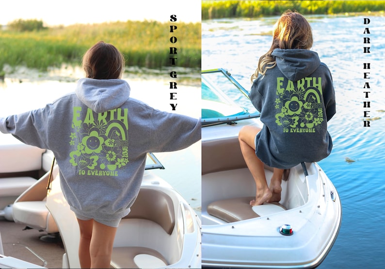 May include: Two women wearing hooded sweatshirts with the text "Earth is Home to Everyone" and a graphic of a smiling flower and rainbow. The sweatshirt on the left is sport grey and the sweatshirt on the right is dark heather grey.