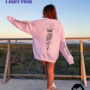May include: A light pink sweatshirt with a black graphic of a crown, a flower, and the word "LOVE" on the back. The sweatshirt has black floral designs on the sleeves.