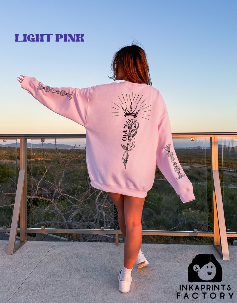May include: A light pink sweatshirt with a black graphic of a crown, a flower, and the word "LOVE" on the back. The sweatshirt has black floral designs on the sleeves.