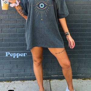 May include: A dark gray oversized t-shirt with a blue and white evil eye graphic design. The graphic features a gold outline and rays of light. The t-shirt is worn by a person standing in front of a brick wall.