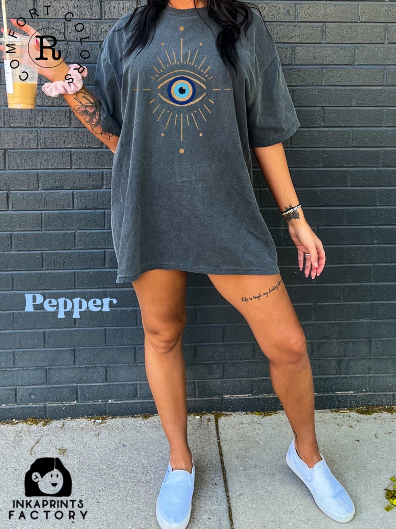 May include: A dark gray oversized t-shirt with a blue and white evil eye graphic design. The graphic features a gold outline and rays of light. The t-shirt is worn by a person standing in front of a brick wall.