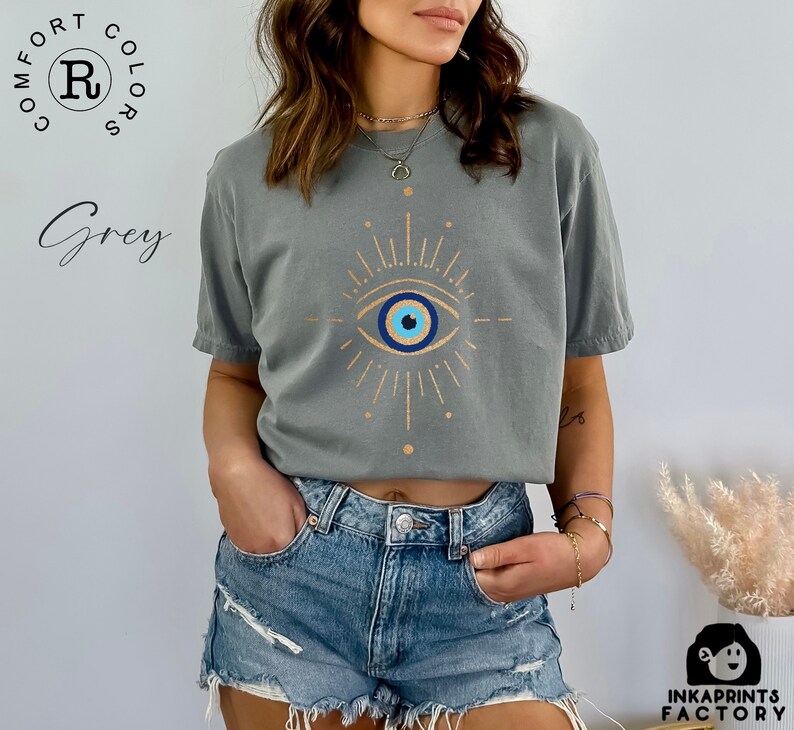 May include: A grey t-shirt with a gold evil eye design. The eye is blue with a white pupil and a black outline. The design is surrounded by gold rays.
