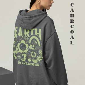 May include: A charcoal gray hooded sweatshirt with a green graphic print on the back. The graphic features the word "Earth" in a retro font, surrounded by flowers, a rainbow, and the words "Peace, Love, To Everyone".