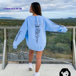 May include: A light blue sweatshirt with a black graphic design on the back. The design features a crown, a flower, and a vine. The sweatshirt has long sleeves and a crew neck.