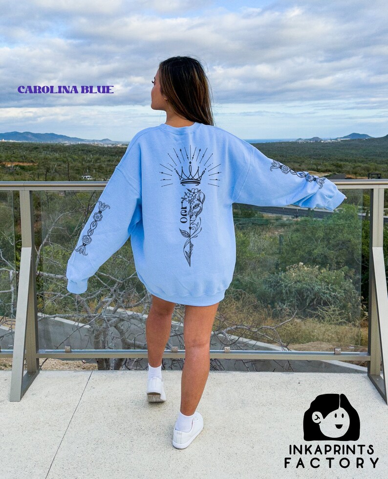 May include: A light blue sweatshirt with a black graphic design on the back. The design features a crown, a flower, and a vine. The sweatshirt has long sleeves and a crew neck.