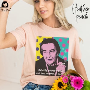 May include: A pink t-shirt with a graphic of a woman with a quote that reads "Authority poisons everybody who takes authority on himself". The graphic is in a pop art style with colorful dots and swirls.