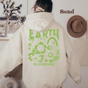 May include: A cream-colored hoodie with a green graphic on the back that says "Earth to Everyone" with a flower, rainbow, and other designs.
