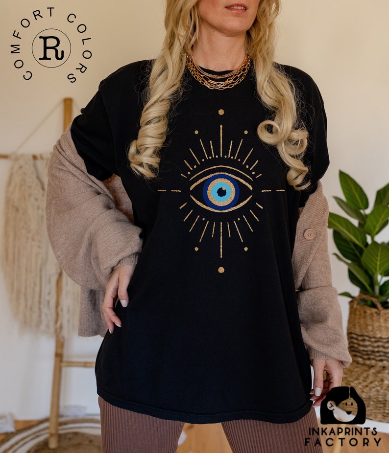 May include: Black t-shirt with a gold evil eye design. The eye is blue with a white pupil and a gold outline. The design is surrounded by gold rays.