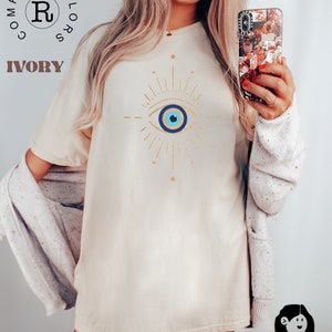 May include: A white t-shirt with a gold evil eye design. The evil eye is blue with a white pupil and a gold outline. The shirt is a relaxed fit and has a crew neck.