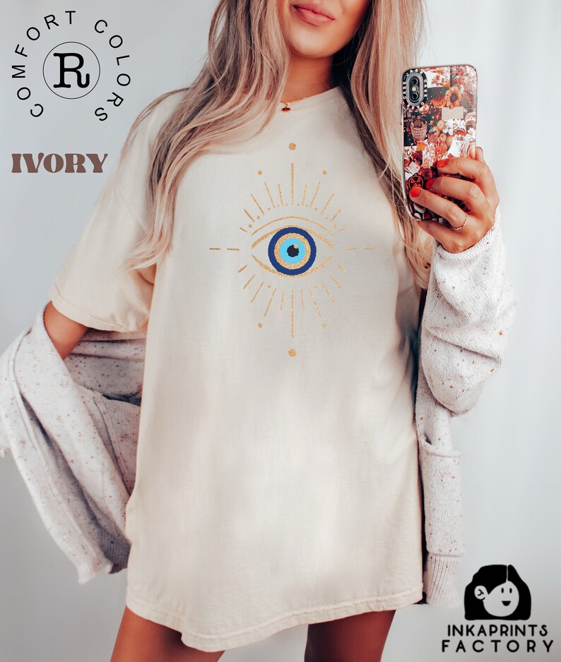 May include: A white t-shirt with a gold evil eye design. The evil eye is blue with a white pupil and a gold outline. The shirt is a relaxed fit and has a crew neck.
